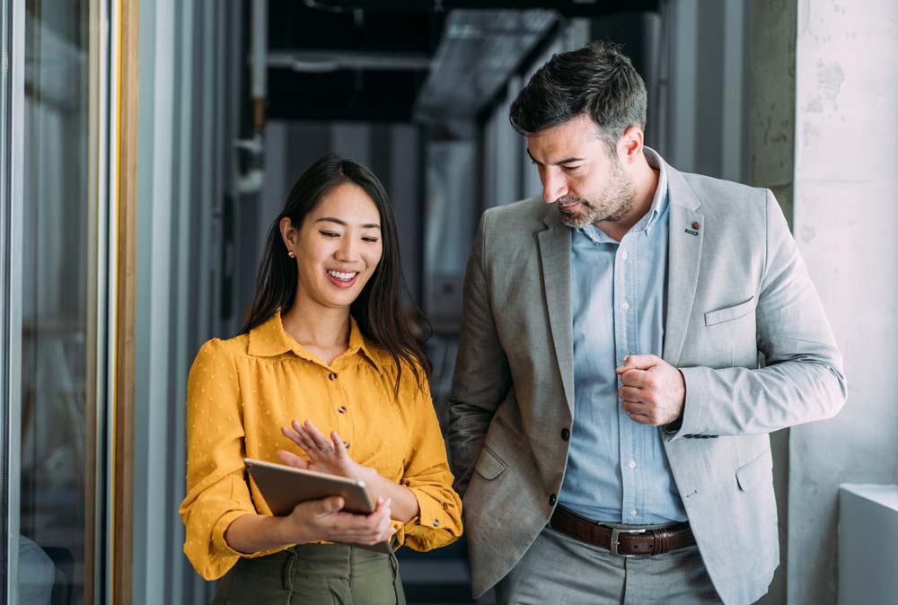 Two colleagues using a digital tablet while walking in a modern office. Businessman and businesswoman in meeting discussing business strategy.